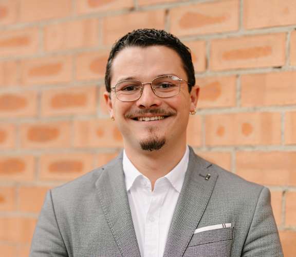 A man in a grey suit stands in front of a brick wall.
