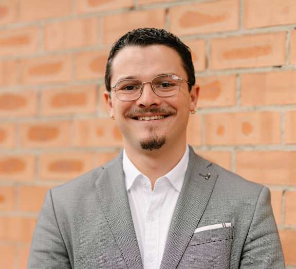A man in a grey suit stands in front of a brick wall.