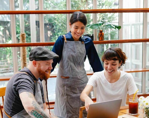 Three people are sitting at a table with a laptop, smiling and enjoying each other's company.