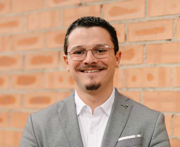 A man in a grey suit stands in front of a brick wall.
