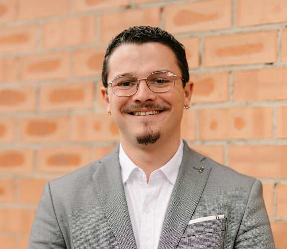 A man in a grey suit stands in front of a brick wall.