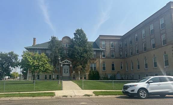A white car is parked in front of The Cornerstone Apartments with a fence and trees.