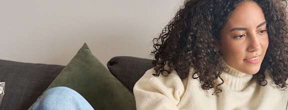 A person with curly hair is sitting on a grey couch using a laptop.