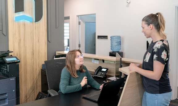 A woman is standing at a reception desk talking to another woman who is seated.