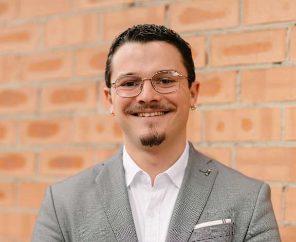 A man in a grey suit stands in front of a brick wall.