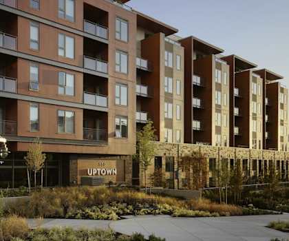 The exterior of the glass, black, and medium brown apartment building showing outdoor balconies and a sidewalk with plants surrounding it during dusk at Uptown at Kirkland Urban. 