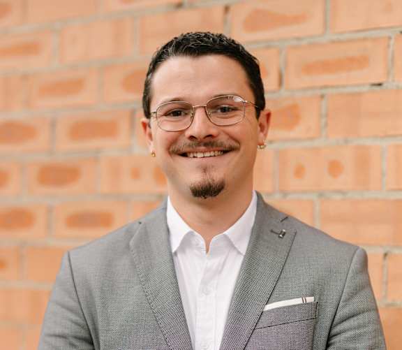 A man in a grey suit stands in front of a brick wall.