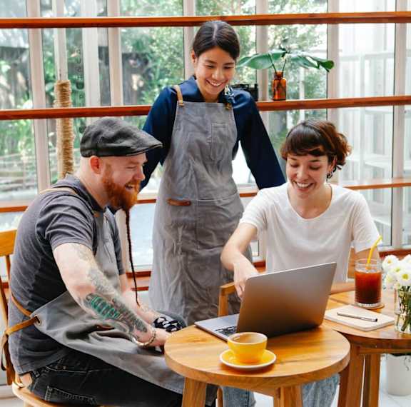 Three people are sitting at a table with a laptop, smiling and enjoying each other's company.