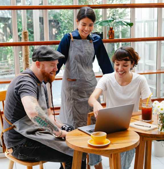 A man with a beard and tattoos is sitting at a table with a laptop, a cup of coffee, and a woman standing behind him.