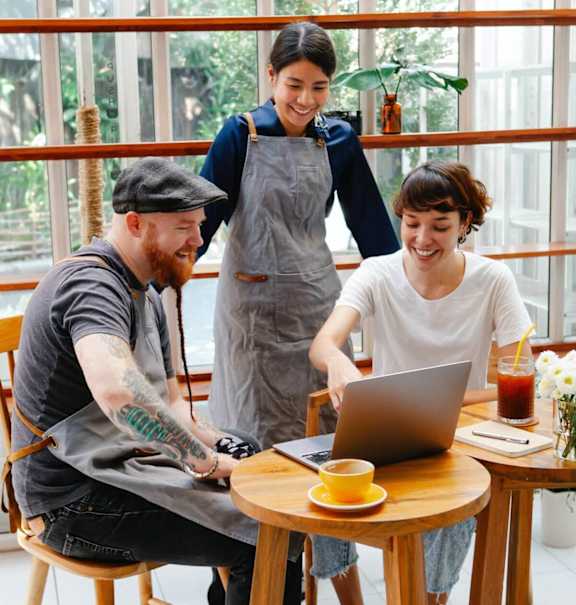 A man with a beard and tattoos is sitting at a table with a laptop, a cup of coffee, and a woman standing behind him.