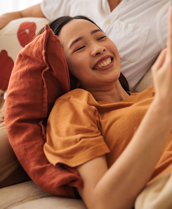 A person is lying on a couch with a pillow, while another person is standing and holding a smartphone.