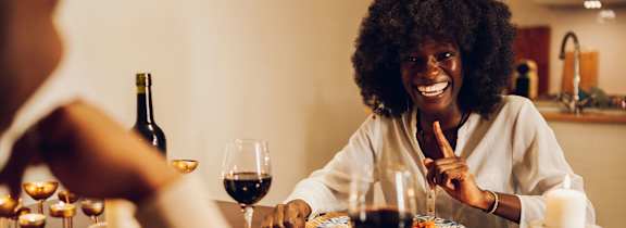 A woman is smiling while sitting at a dinner table with a glass of wine in front of her.