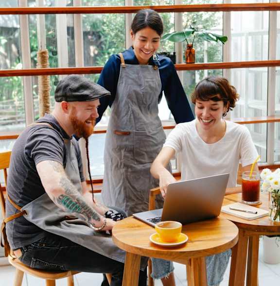 A man with a beard and tattoos is sitting at a table with a laptop, a cup of coffee, and a woman standing behind him.