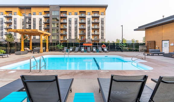 A large swimming pool with lounge chairs and a wooden pergola in front of a multi-story apartment building.