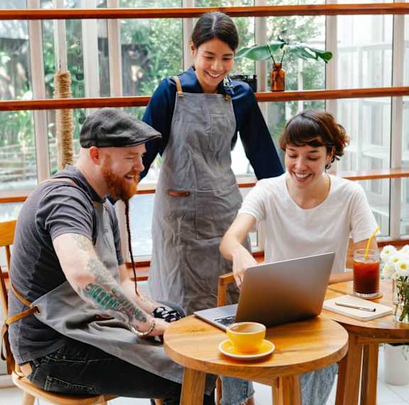A man with a beard and tattoos is sitting at a table with a laptop, a cup of coffee, and a woman standing behind him.