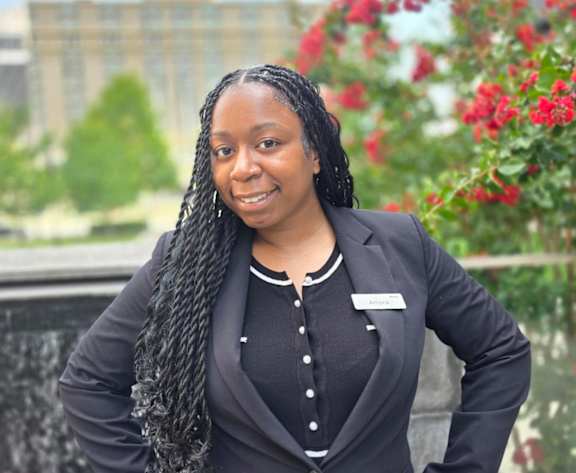 A woman with braided hair wearing a black dress and a name tag is standing in front of a fountain.