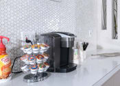 Thumbnail 18 of 23 - Fifteen 50 apartments Las Vegas clubhouse coffee corner with Keurig and coffee cups on counter with modern white penny tile backsplash.