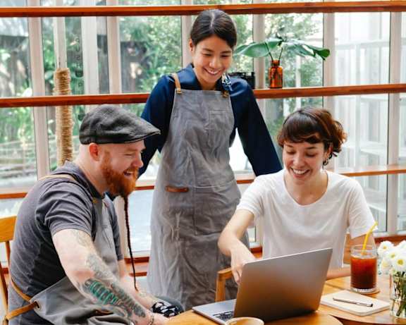 A man with a beard and tattoos is sitting at a table with a laptop, a cup of coffee, and a woman standing behind him.