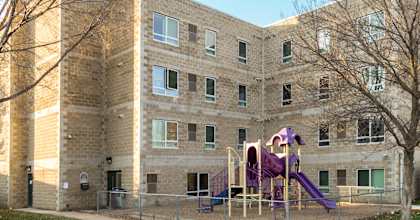 a 4 story tall tan brick building with a playground in the forefront with a purple slide, tunnel, and roof
