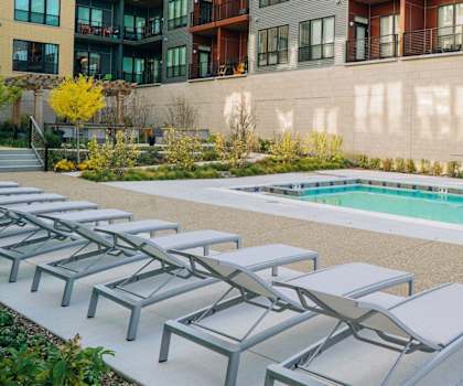 The large, turquoise outdoor pool with pool chairs, tables, and chairs with the apartments in the background at River House Apartment Homes. 