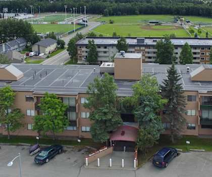 The arial view of the brown apartment buildings surrounded by trees with the resident parking lot in the foreground and the neighboring apartment buildings and baseball field in the background at the Outlook Apartment Homes. 