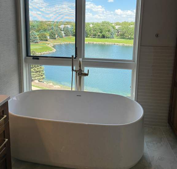 A white bathtub is in a bathroom with a window overlooking a large pond