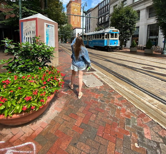 A woman in a denim jacket and shorts stands on a brick walkway next to a flower bed and a tram.