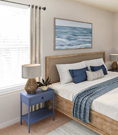 Bright apartment bedroom with wood bed frame, blue nightstand, and large windows at Palmetto Place Apartments in Taylors, SC