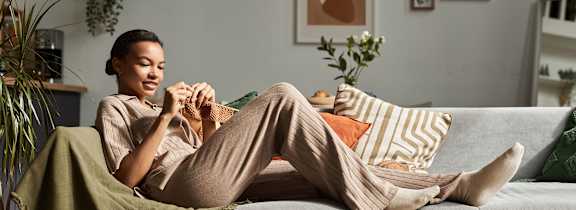 A woman is sitting on a couch with a green blanket, holding a book and a cup of coffee.