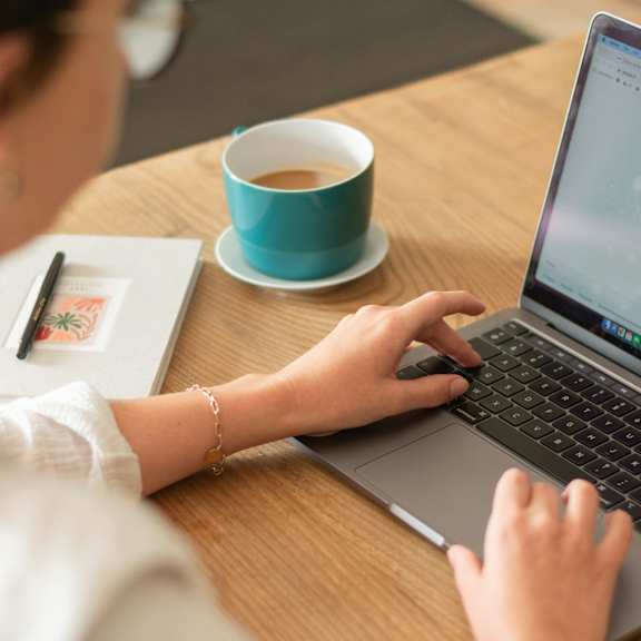 A person is typing on a laptop with a cup of coffee and a notebook beside them.