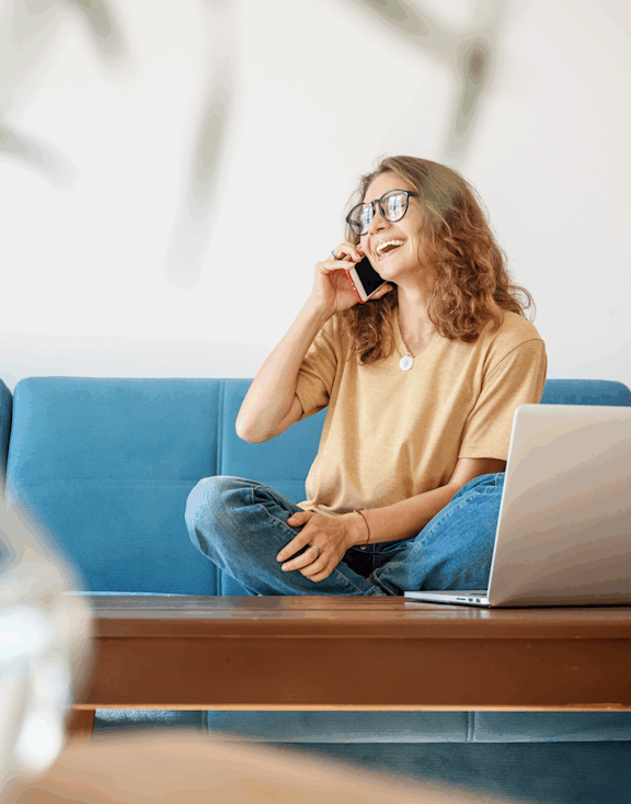A woman with glasses is talking on the phone while sitting on a blue couch with a laptop on her lap.