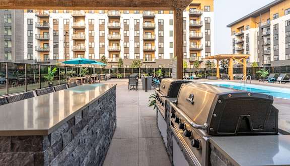 A large outdoor grill is in the foreground of a patio with a pool and apartment buildings in the background.