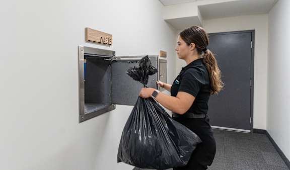 A woman is holding a black trash bag in front of a trash can  at Vibe Apartments at Arbour Lake in Calgary, AB