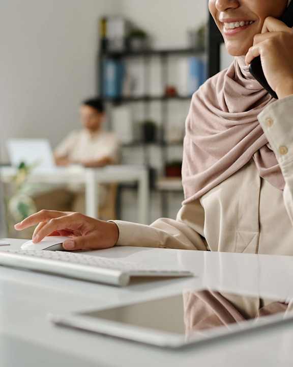 A woman in a hijab is talking on the phone while working on her computer.