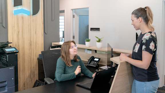 A woman is standing at a reception desk talking to another woman who is seated.