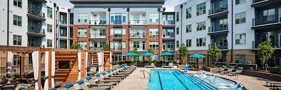 A large swimming pool surrounded by lounge chairs and umbrellas in front of apartment buildings.