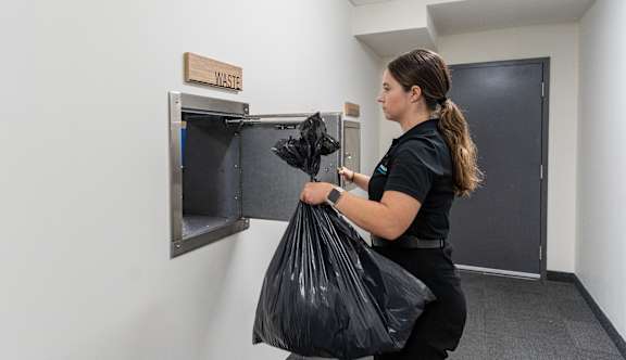 A woman is holding a black trash bag in front of a trash can.