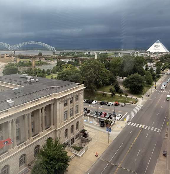 A view of a city street with a large building on the left and a bridge in the distance under a cloudy sky.
