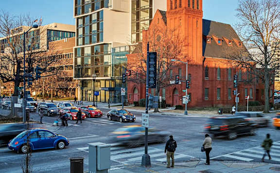 A busy street scene with cars and pedestrians in front of a modern building and a historic church.
