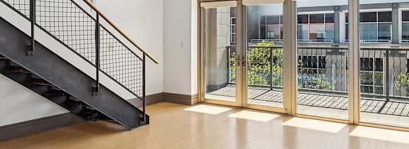 A room with a wooden floor, a staircase, and a view of the outside through the windows.