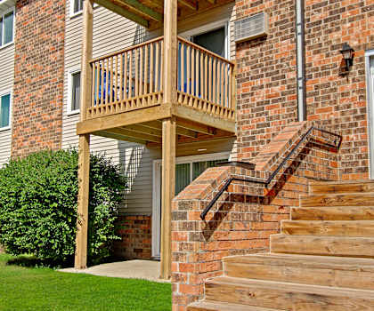 The red brick front entrance to one of the apartment buildings showing a balcony, patio, windows, and a grassy area with blue sky above at Windscape Apartment Homes. 