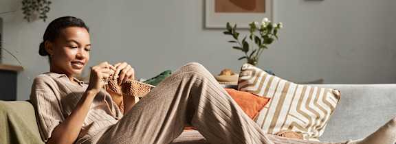 A woman is sitting on a couch with a green blanket, holding a book and a cup of coffee.