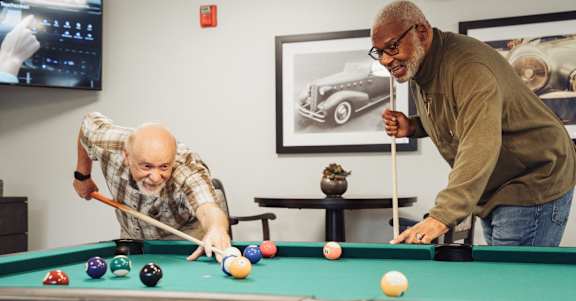 Two men playing pool in a room with a TV and a vintage car poster on the wall.