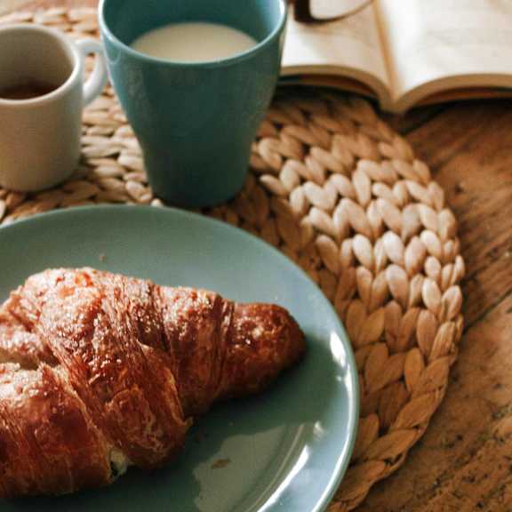 A blue plate with a croissant and a blue cup of milk next to an open book.