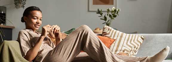 A woman is sitting on a couch with a green blanket, holding a book and a cup of coffee.