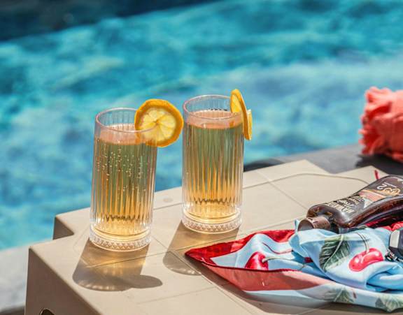 Two glasses of a golden-colored beverage with a slice of orange on the rim, placed on a table beside a pool.