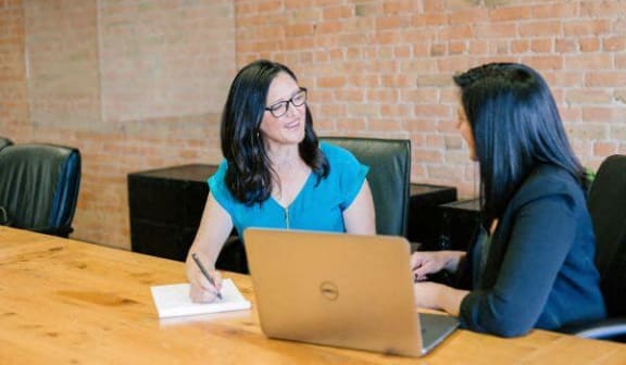 two women sitting at a table with a laptop