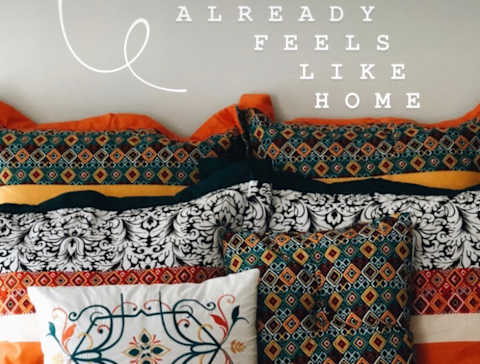 A bed with a colorful comforter and pillows with the text "ALREADY FEELS LIKE HOME" overlaid.