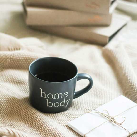 A black mug with the words 'home body' next to a stack of books.