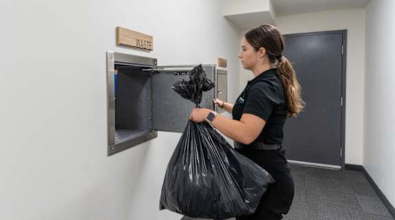 A woman is holding a black trash bag in front of a trash can.